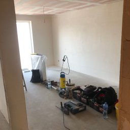 S-BAU USŁUGI OGÓLNOBUDOWLANE STANKIEWICZ - Pokój w trakcie remontu: widoczne płyty karton gips na suficie, częściowo zaszpachlowane, oraz narzędzia i materiały budowlane rozłożone na podłodze, w tym wiertarka, opryskiwacz i pojemniki...