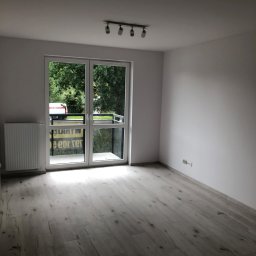 S-BAU USŁUGI OGÓLNOBUDOWLANE STANKIEWICZ - Puste, jasne pomieszczenie z panelami podłogowymi w odcieniach szarości, grzejnikiem pod oknem balkonowym i listwą oświetleniową na suficie.