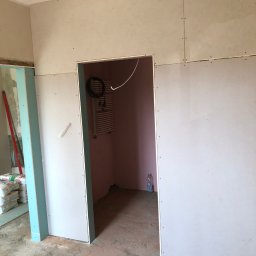S-BAU USŁUGI OGÓLNOBUDOWLANE STANKIEWICZ - Montaż płyt karton gips na ścianie, widoczny otwór drzwiowy, w tle grzejnik drabinkowy i butelka wody, na podłodze ślady po zaprawie, worki z zaprawą Cekol w lewym dolnym rogu.