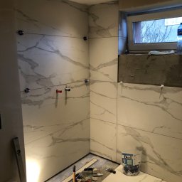 S-BAU USŁUGI OGÓLNOBUDOWLANE STANKIEWICZ - Łazienka w trakcie montażu płytek imitujących marmur, widoczne krzyżyki dystansowe, poziomica i narzędzia glazurnicze oraz częściowo wykończone obramowanie okna.