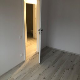 S-BAU USŁUGI OGÓLNOBUDOWLANE STANKIEWICZ - Pusty pokój z nową, jasną podłogą imitującą drewno, otwarte białe drzwi z czarną klamką prowadzą do łazienki, widoczny fragment toalety.