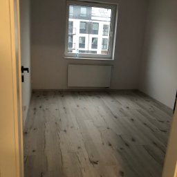 S-BAU USŁUGI OGÓLNOBUDOWLANE STANKIEWICZ - Pusty pokój z panelami podłogowymi w odcieniach szarości i bieli, widok zza uchylonych drzwi, okno z widokiem na budynek mieszkalny w tle.