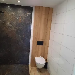 Nowoczesna łazienka z marmurową ścianą prysznicową, drewnianym panelem z czarnym przyciskiem spłuczki i białymi płytkami ściennymi.