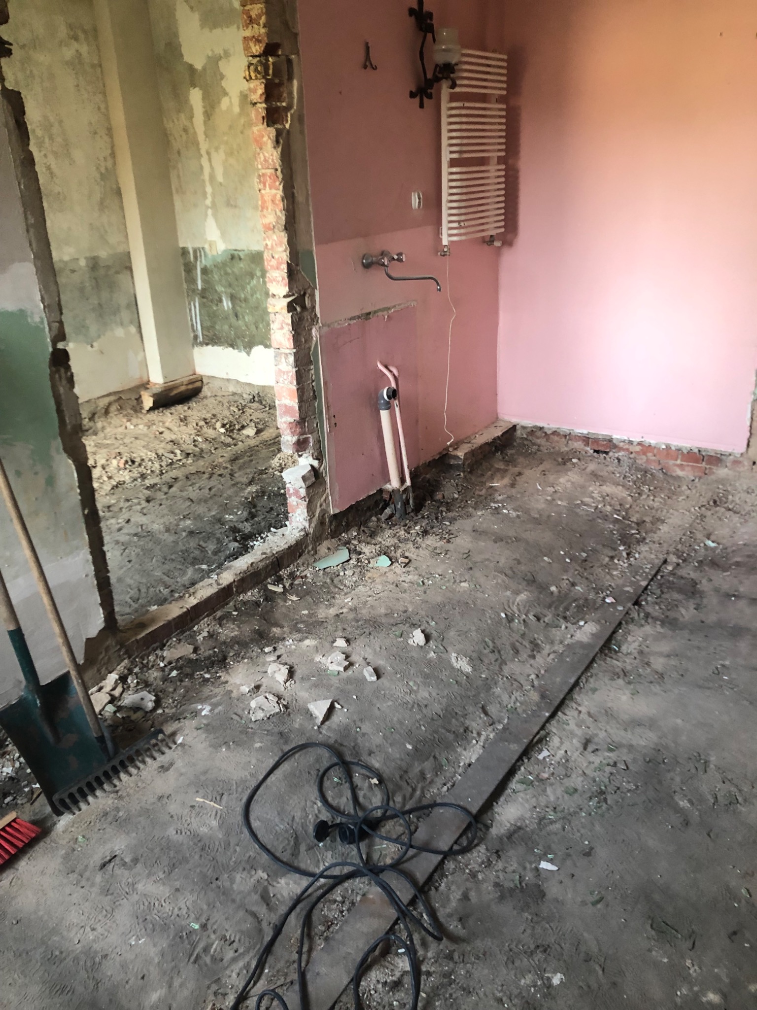 Łazienka w trakcie demontażu: różowe ściany, zdemontowane płytki, widoczne rury, grzejnik na ścianie, podłoga pokryta gruzem i ziemią, narzędzia w rogu.