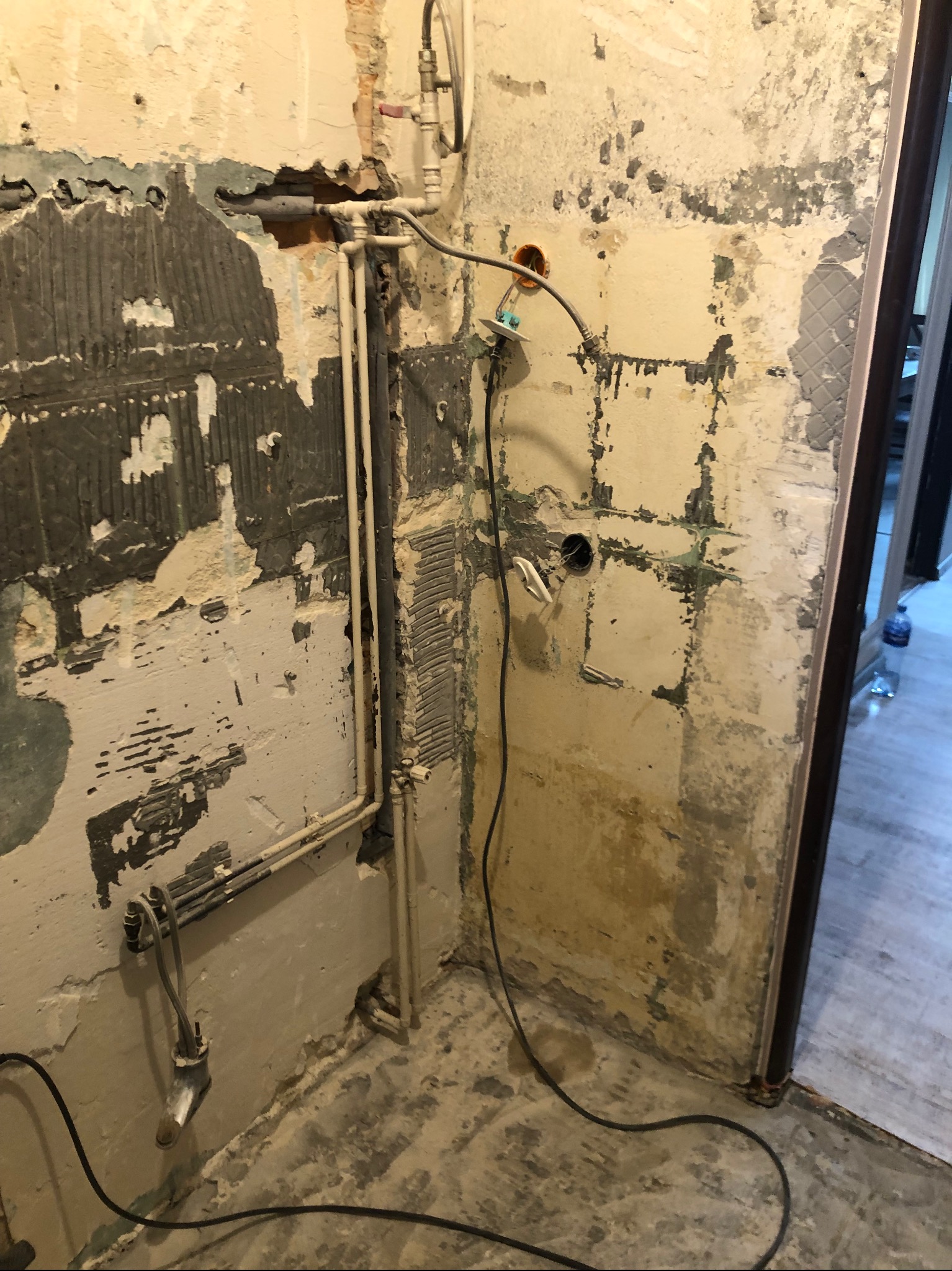 Fragment łazienki w trakcie demontażu starych płytek, z widocznymi instalacjami hydraulicznymi i elektrycznymi oraz resztkami kleju na ścianach.