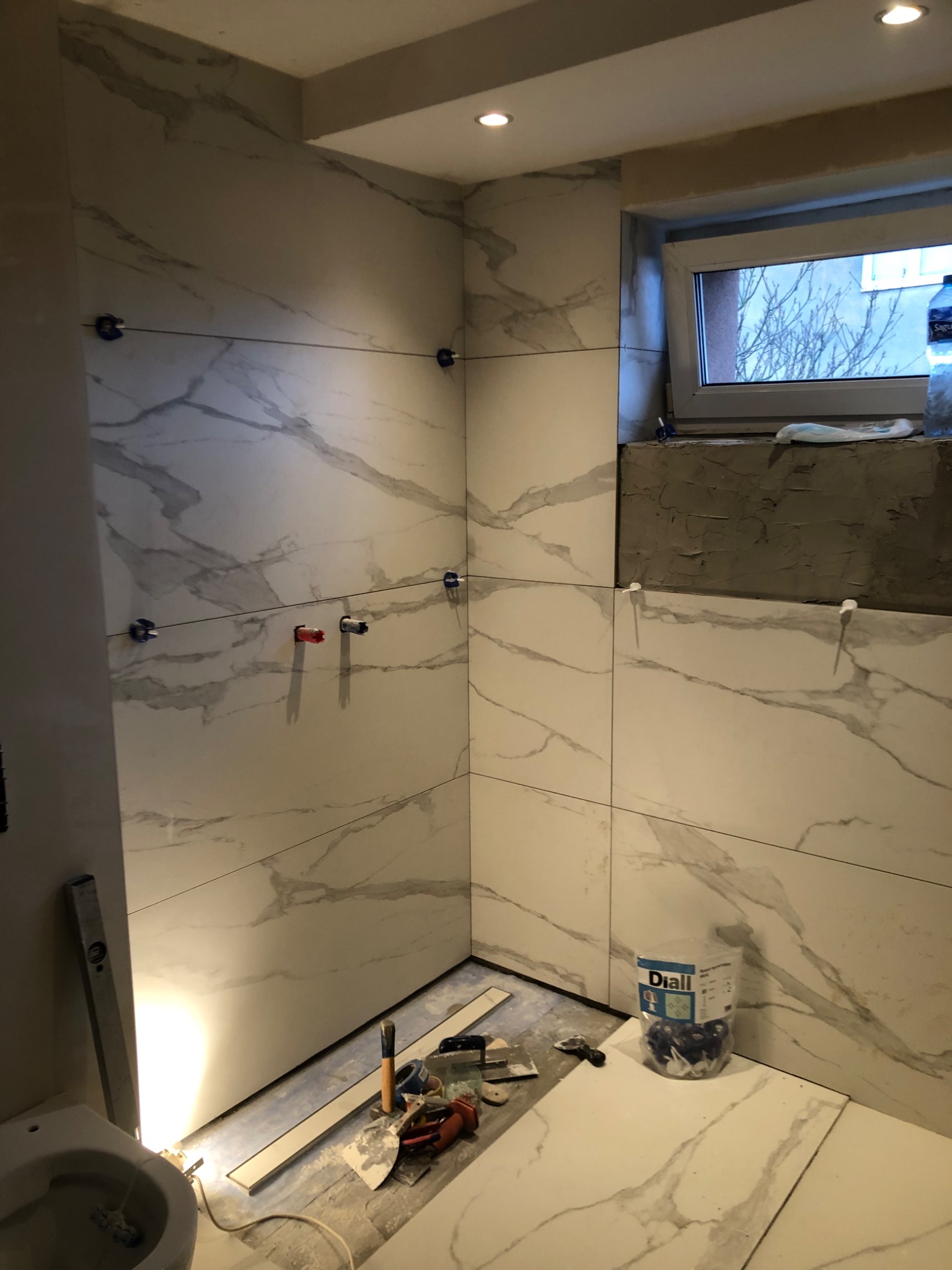 Łazienka w trakcie montażu płytek imitujących marmur, widoczne krzyżyki dystansowe, poziomica i narzędzia glazurnicze oraz częściowo wykończone obramowanie okna.