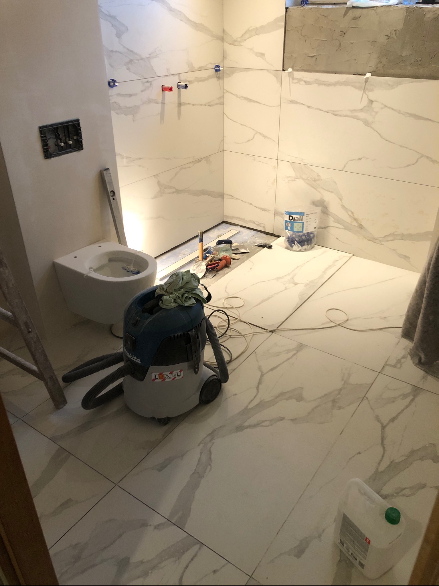 Montaż płytek w łazience w trakcie realizacji, widoczny podwieszany sedes, odkurzacz budowlany Makita, poziomica, krzyżyki glazurnicze Diall i częściowo ułożona podłoga z płytek imitujących marmur.