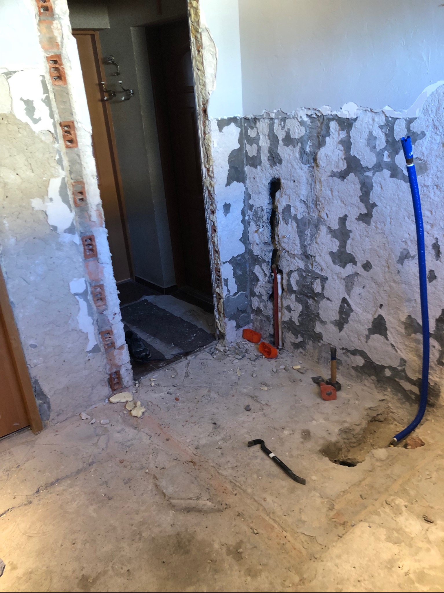 Fragment pomieszczenia w trakcie prac przygotowawczych do remontu: ogołocone ściany z resztkami tynku, widoczne instalacje hydrauliczne, fragment otworu drzwiowego z odsłoniętą cegłą, na podłodze...