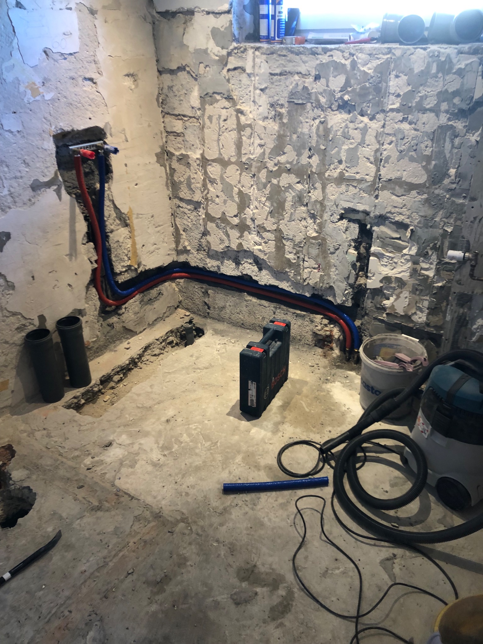 Surowa ściana łazienki w trakcie prac instalacyjnych z widocznymi rurami ciepłej i zimnej wody, narzędziami w kufrze Bosch oraz odkurzaczem przemysłowym.