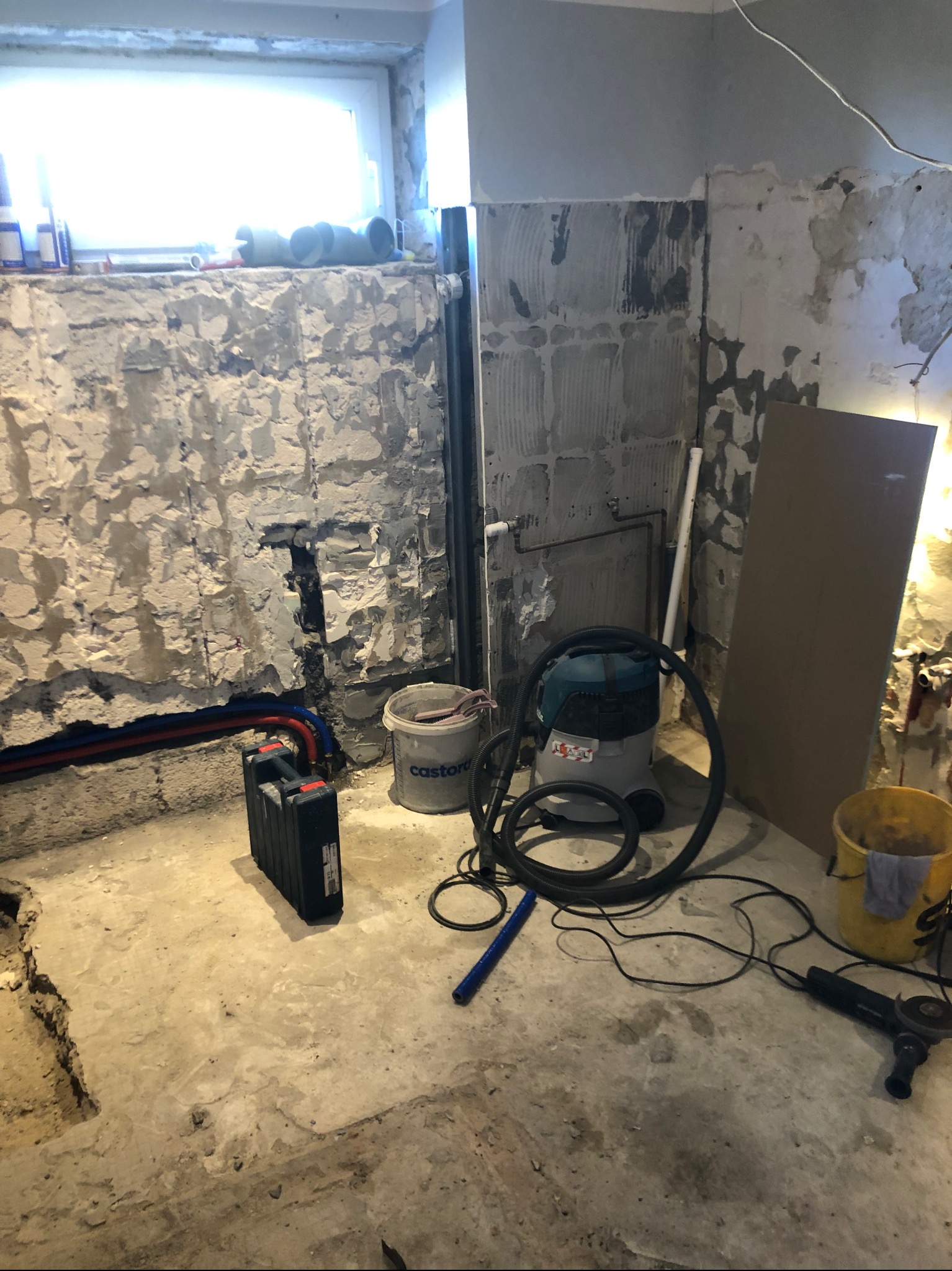 Wnętrze w trakcie prac przygotowawczych: odsłonięte mury z instalacją, widoczny odkurzacz przemysłowy, wiadro i szlifierka kątowa na surowym podłożu.