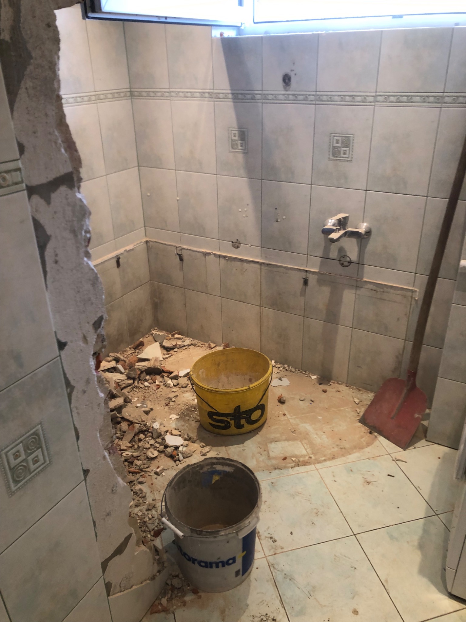 Demontaż płytek w łazience, widoczne gruz, wiadro z napisem STO, odsłonięte fragmenty ściany i podłogi, przygotowanie do remontu.