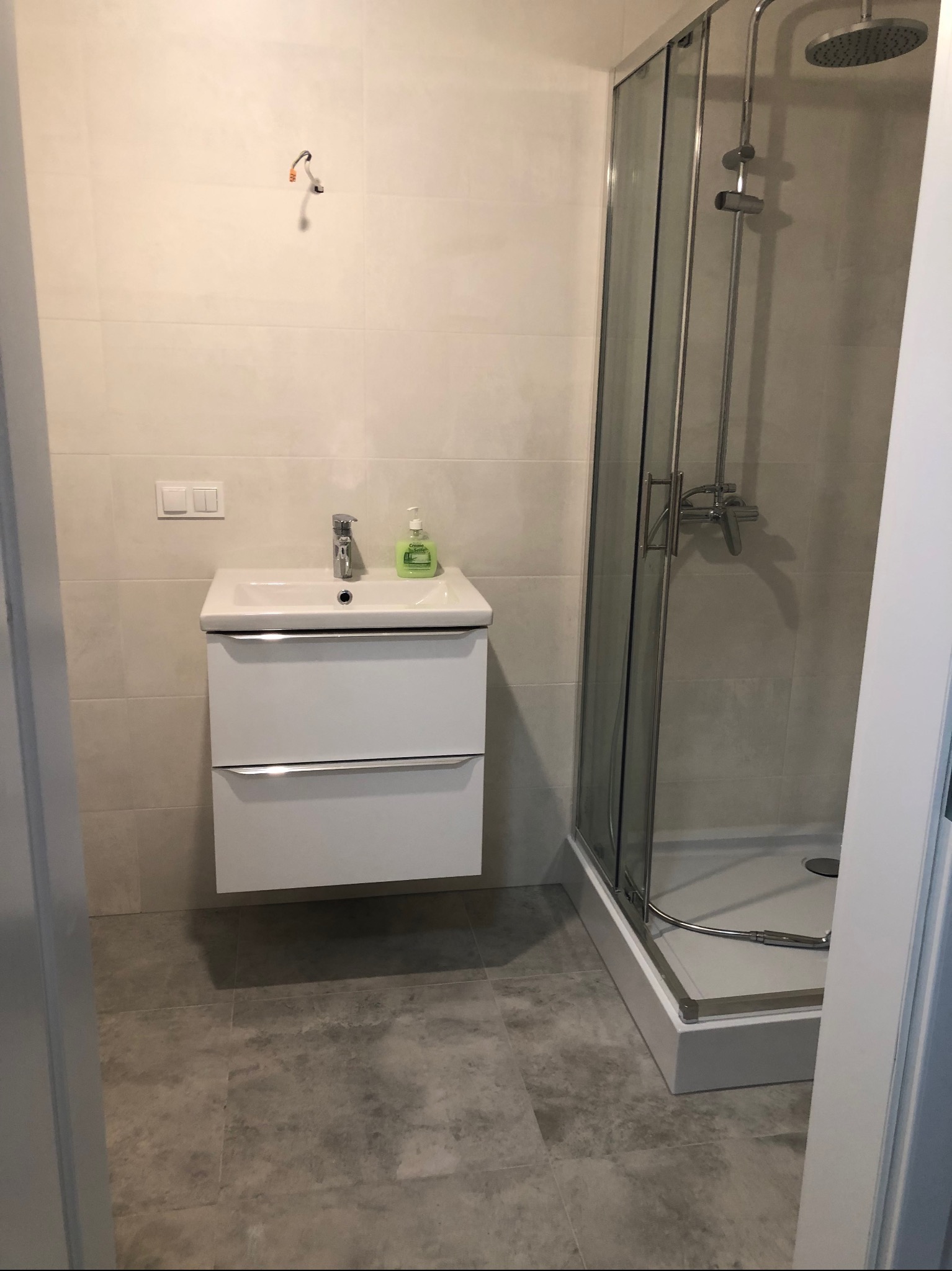 Jasna łazienka po remoncie z kabiną prysznicową, umywalką z szafką i szarą podłogą; widoczne niedokończone podłączenie elektryczne na ścianie.