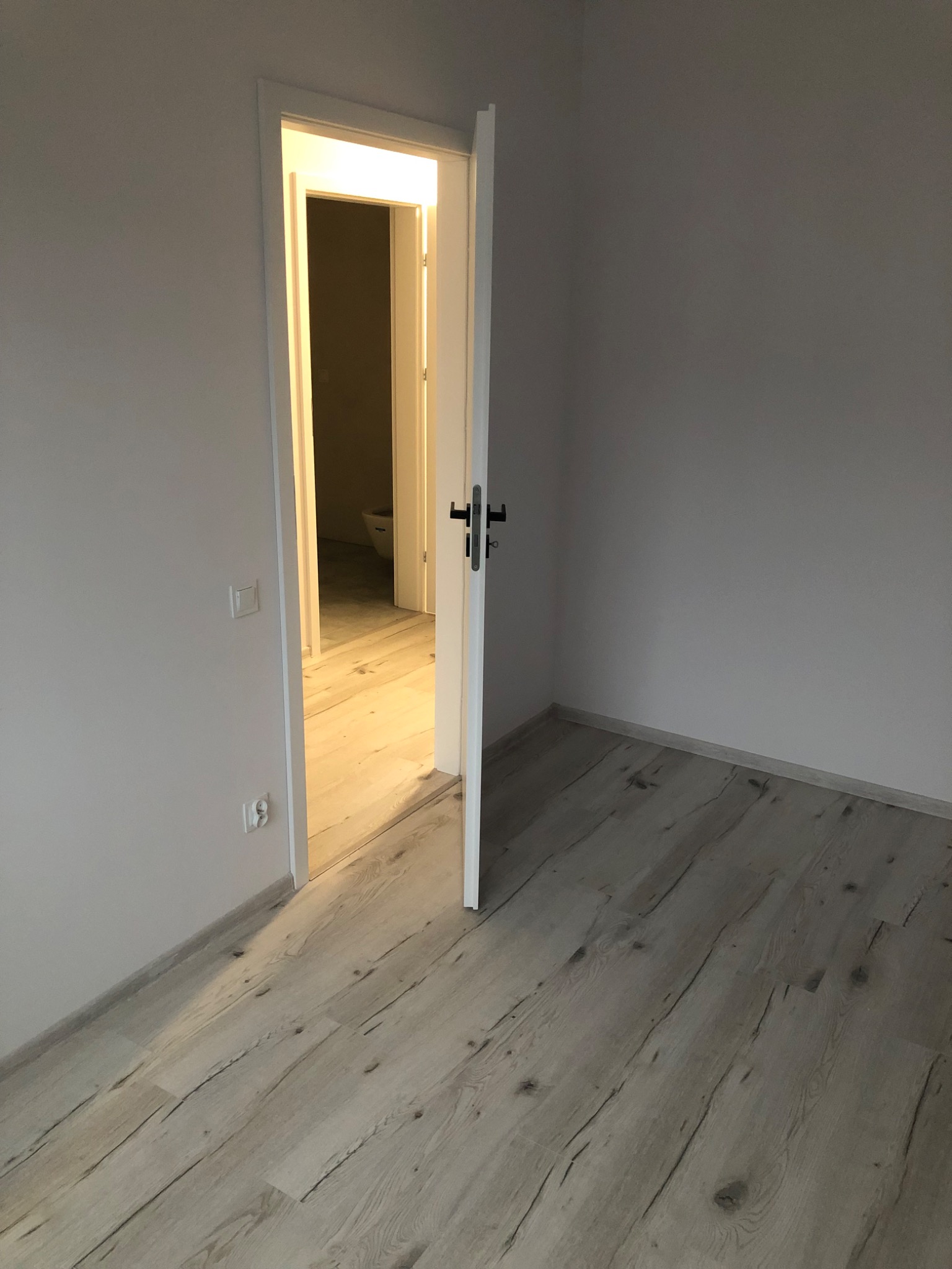 Pusty pokój z nową, jasną podłogą imitującą drewno, otwarte białe drzwi z czarną klamką prowadzą do łazienki, widoczny fragment toalety.