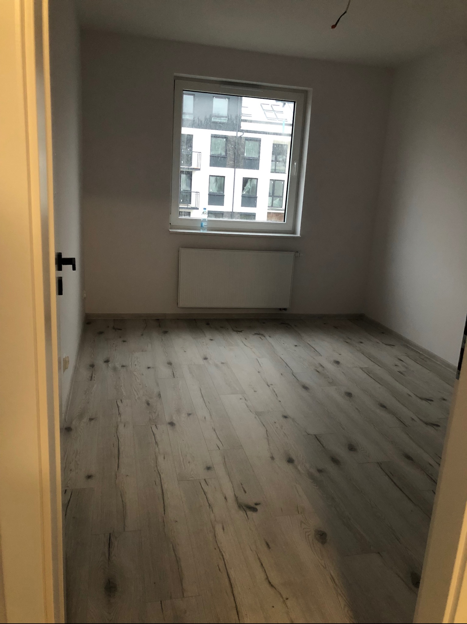 Pusty pokój z panelami podłogowymi w odcieniach szarości i bieli, widok zza uchylonych drzwi, okno z widokiem na budynek mieszkalny w tle.