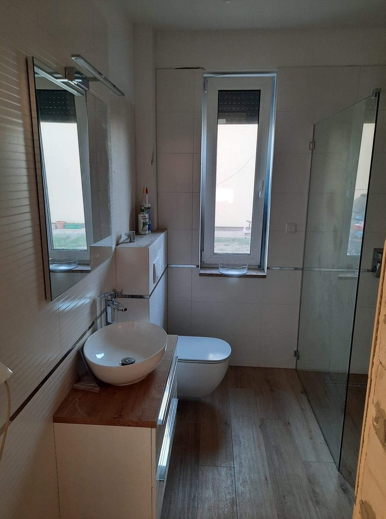 Wnętrze łazienki w trakcie wykańczania, widoczna umywalka nablatowa na drewnianym blacie, toaleta wisząca, kabina prysznicowa ze szklaną ścianą oraz okno z roletą, płytki na ścianach i podłodze...