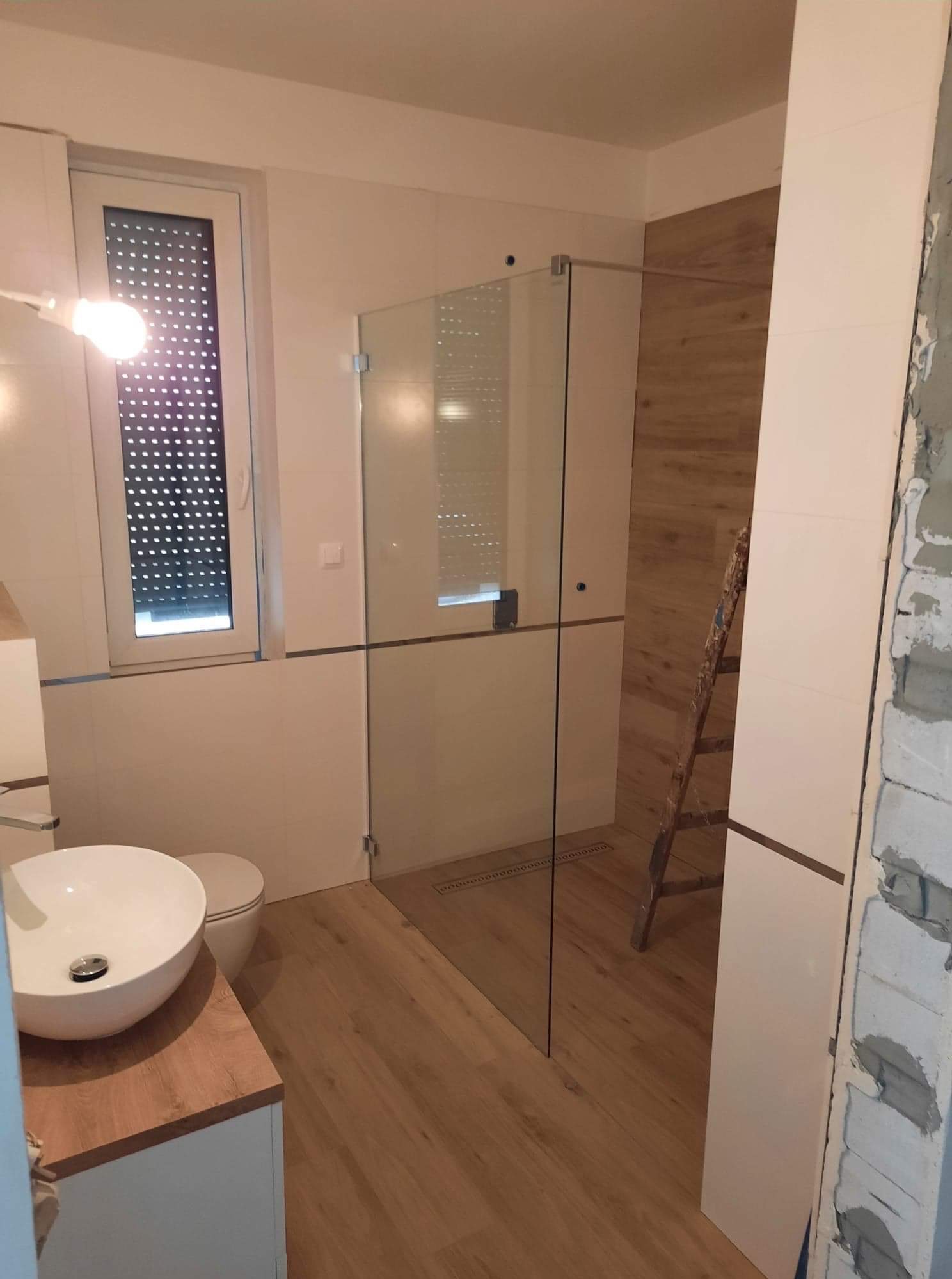 Nowoczesna łazienka w trakcie wykańczania: kabina prysznicowa ze szklanymi drzwiami, umywalka nablatowa na drewnianym blacie, drewnopodobne płytki na podłodze i ścianie pod prysznicem, drabina...