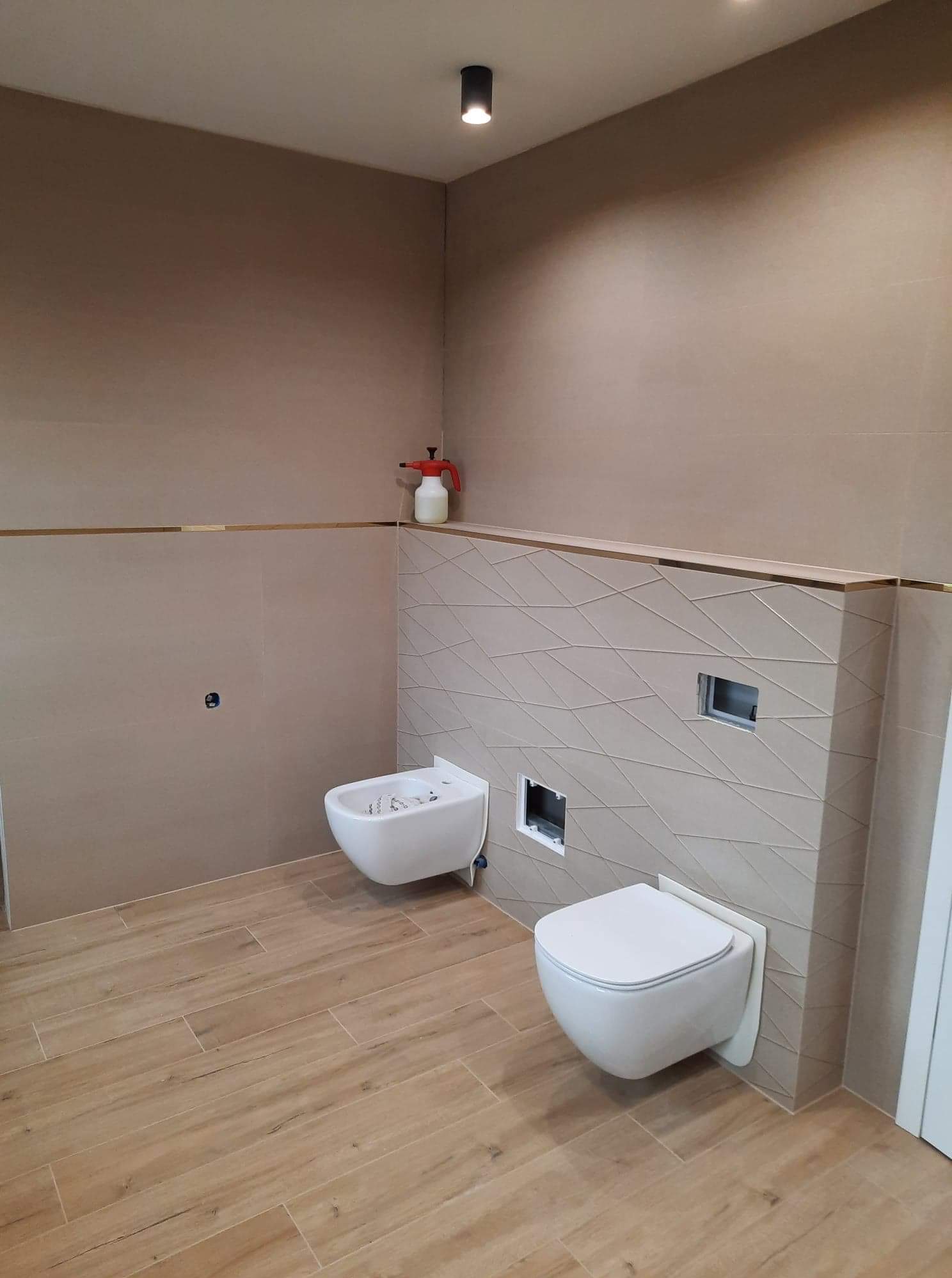 Nowoczesna łazienka w trakcie montażu armatury: wiszący bidet i toaleta, beżowe płytki z dekoracyjnym wzorem na ścianie, drewnopodobna podłoga, widoczny spryskiwacz na półce.