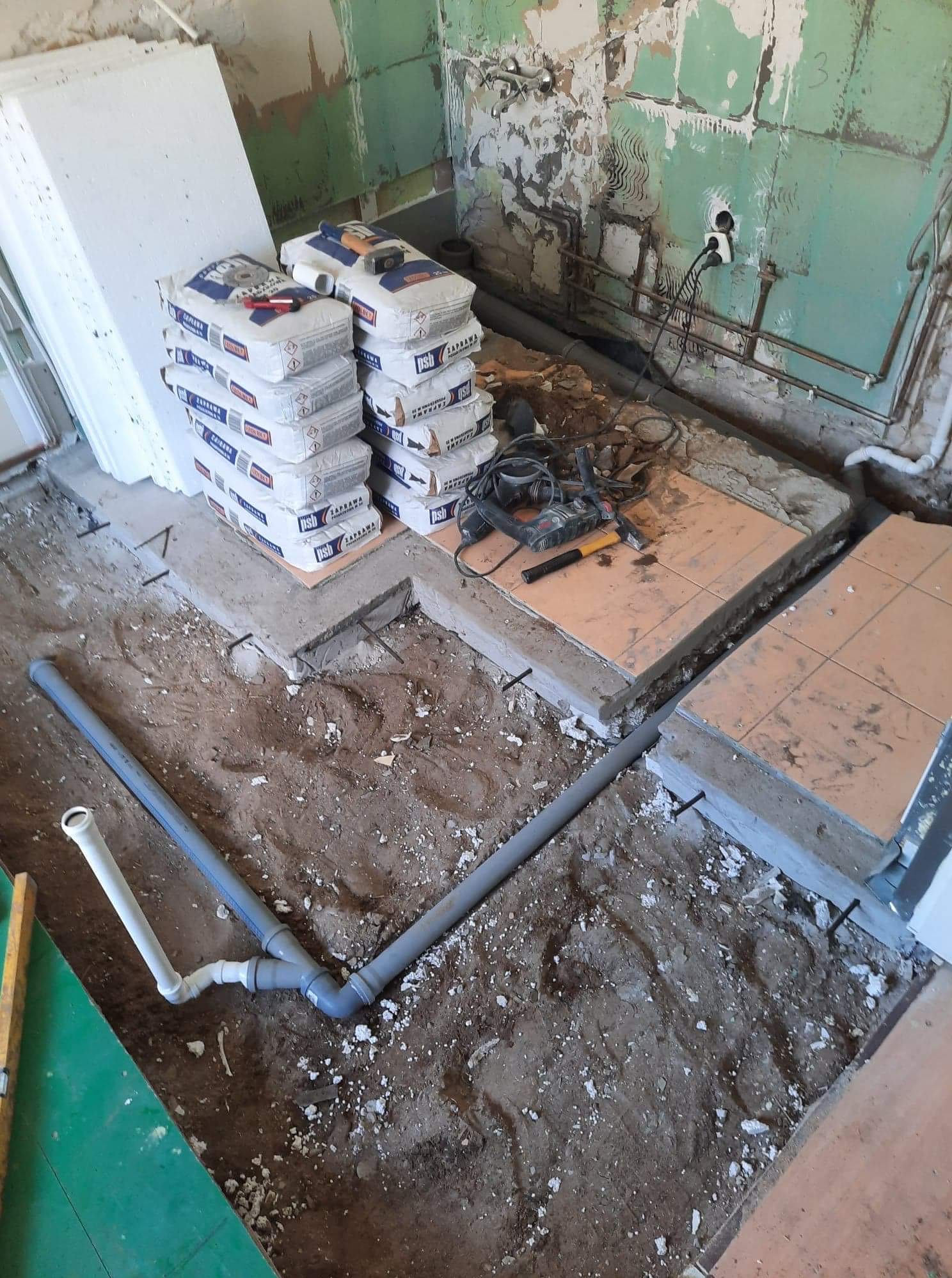 Fragment łazienki w trakcie prac: ułożone rury kanalizacyjne w wykopie, obok worki z zaprawą, płyty styropianowe i narzędzia na podwyższonej części podłogi z fragmentami płytek.