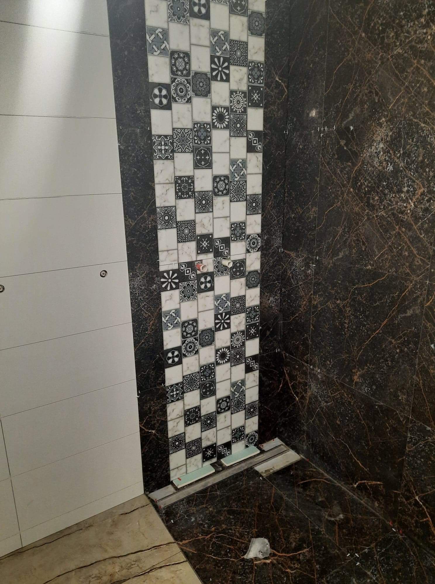 Montaż płytek w łazience: wzorzyste płytki dekoracyjne na ścianie, kontrastujące z ciemnym marmurem i białymi płytami, widoczne krzyżyki dystansowe.