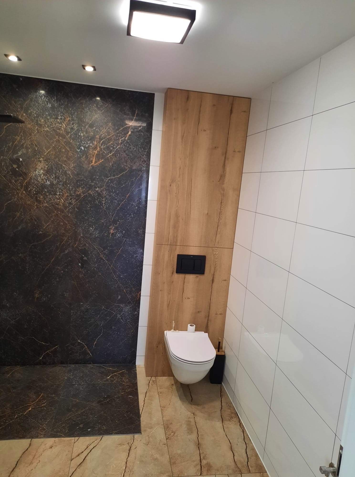 Nowoczesna łazienka z marmurową ścianą prysznicową, drewnianym panelem z czarnym przyciskiem spłuczki i białymi płytkami ściennymi.