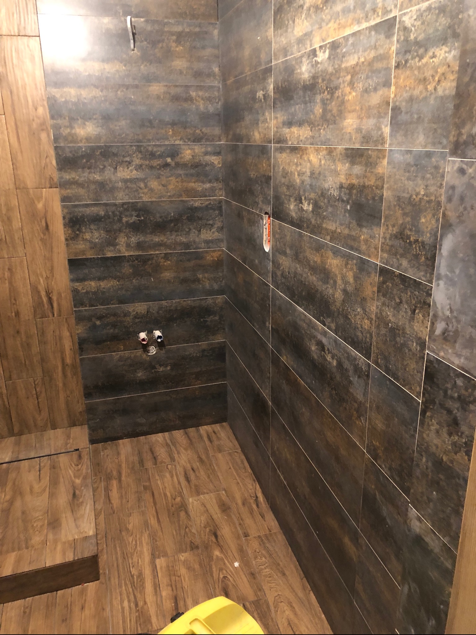 Nowoczesna kabina prysznicowa z ciemnymi, metalicznymi płytkami imitującymi rdzę i drewnopodobną podłogą, widoczne przyłącza wodne i fragment żółtego urządzenia czyszczącego.