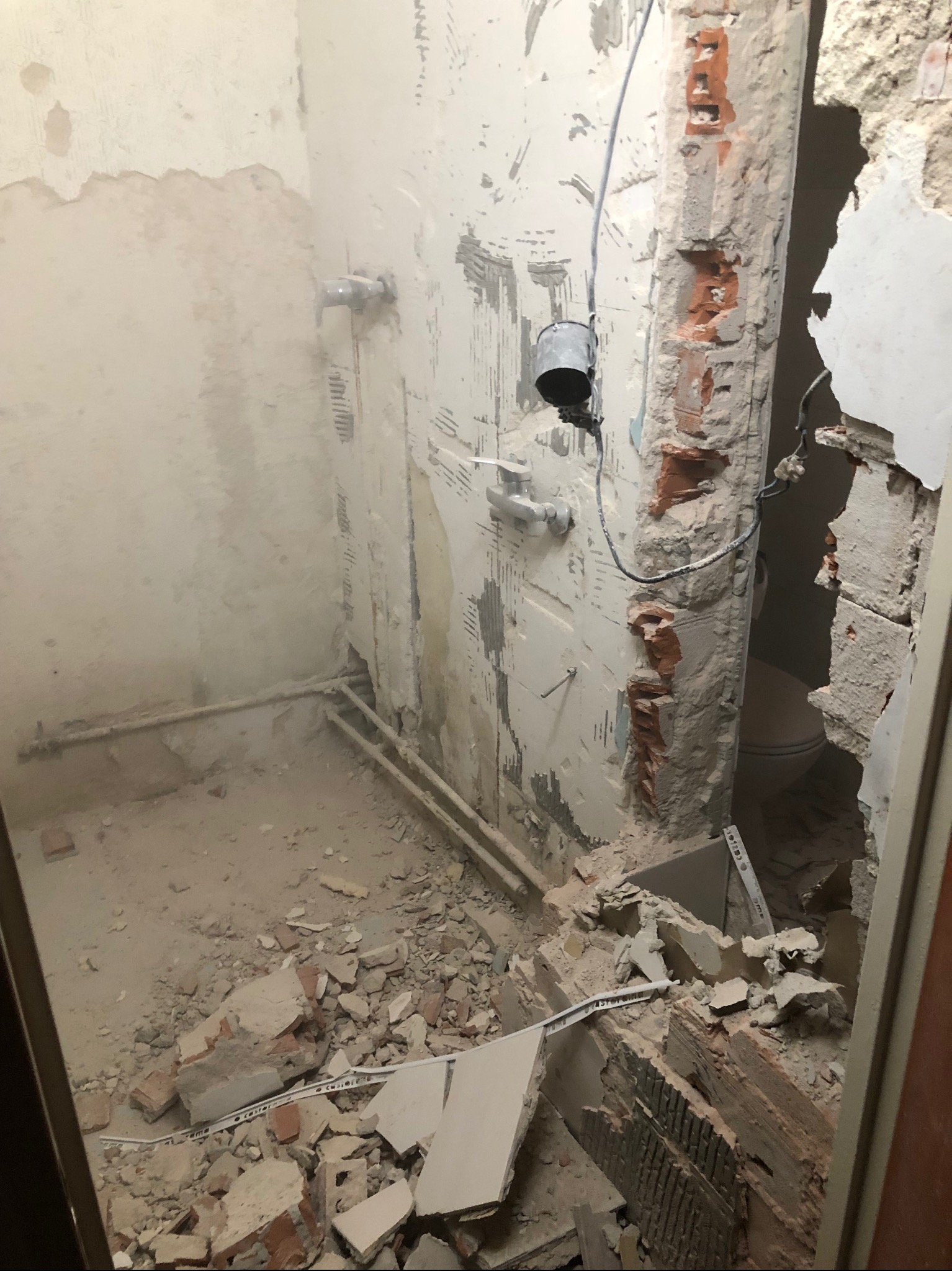 Łazienka w trakcie generalnego remontu: zdemontowane płytki, gruz na podłodze, odsłonięte rury i fragmenty cegieł w ościeżnicy