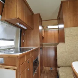 Wnętrze kampera z zabudową drewnianą: aneks kuchenny z płytą grzewczą, szafkami i oknem z roletą, obok tapicerowane siedzisko z pasami bezpieczeństwa i stolik, widoczna również część sypialna...