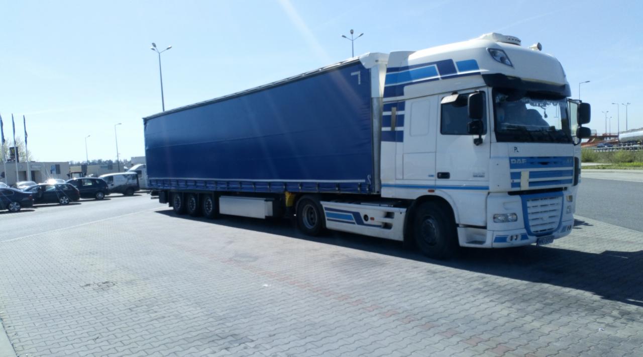 Biało-niebieska ciężarówka DAF z plandeką, zaparkowana na placu wyłożonym kostką brukową, z widocznymi samochodami osobowymi w tle i numerem '7' na plandece.