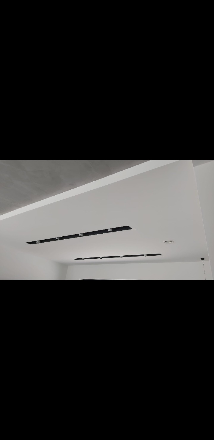 Biały sufit z dwoma rzędami wpuszczanych, czarnych lamp. Widoczne gładkie wykończenie i minimalistyczny design. Oświetlenie punktowe.