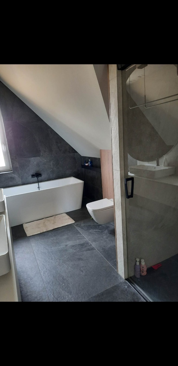 Nowoczesna łazienka na poddaszu z wanną, toaletą i umywalką. Ciemne płytki podłogowe i ścienne kontrastują z bielą armatury. Widoczne okno dachowe i kabina prysznicowa.