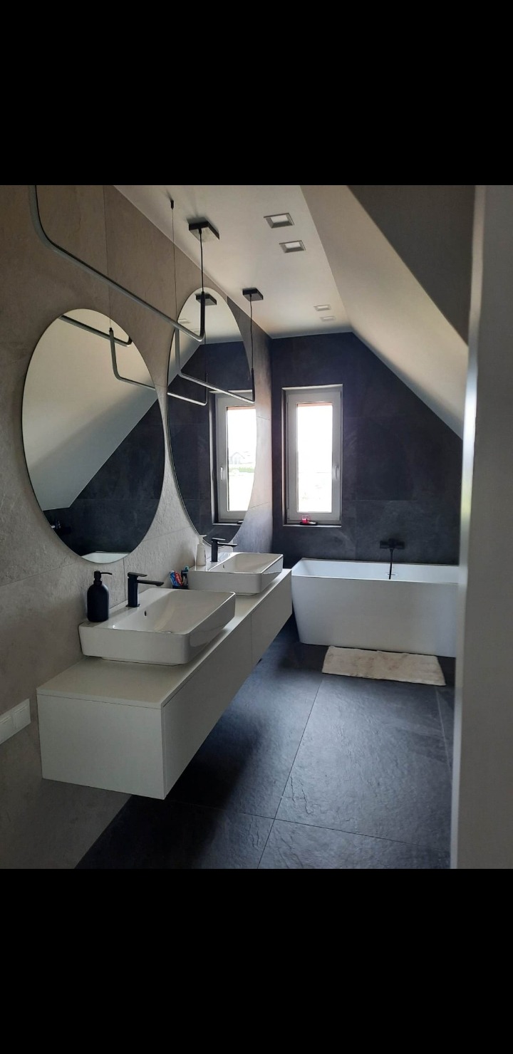 Nowoczesna łazienka na poddaszu z dwoma okrągłymi lustrami, białą szafką pod umywalki i wanną. Ciemne płytki na podłodze i ścianach, minimalistyczne oświetlenie.