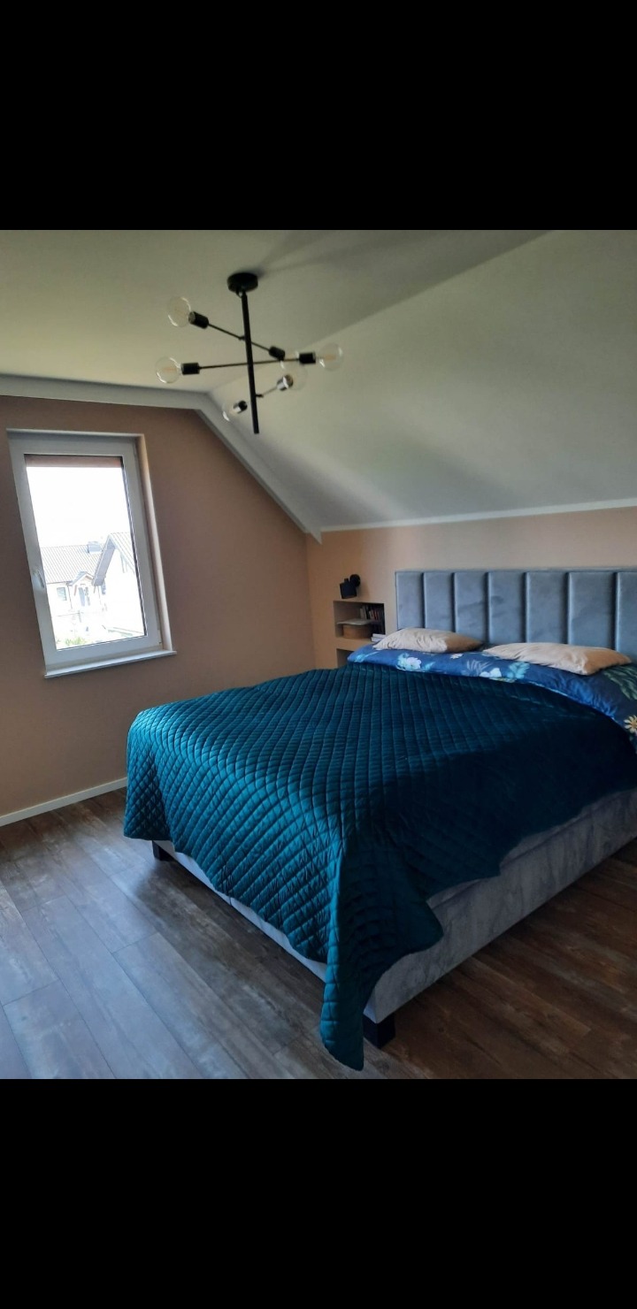 Sypialnia na poddaszu z łóżkiem w szaro-turkusowej tonacji, widok na okno i nowoczesną lampę sufitową. Ściany w odcieniach beżu.