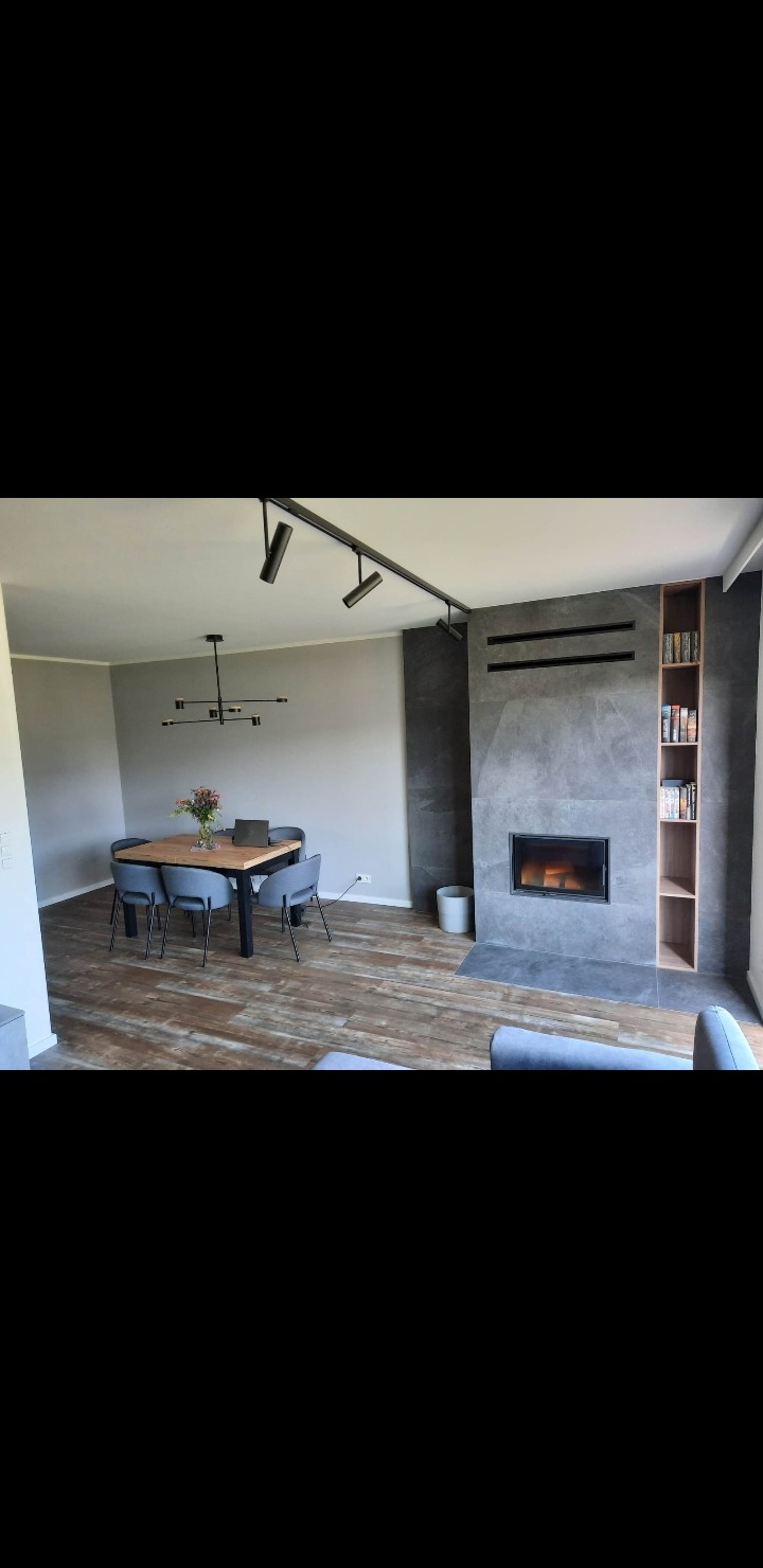 Nowoczesny salon z szarymi ścianami, drewnianą podłogą i kominkiem obłożonym szarą płytą. Nad stołem designerska lampa, a nad kominkiem półki na książki.