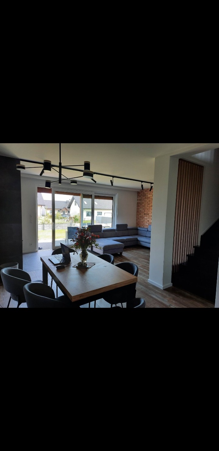 Nowoczesne wnętrze domu z drewnianym stołem, szarą sofą narożną i ceglaną ścianą. Widoczne schody i wyjście na ogród. Czarna lampa sufitowa.