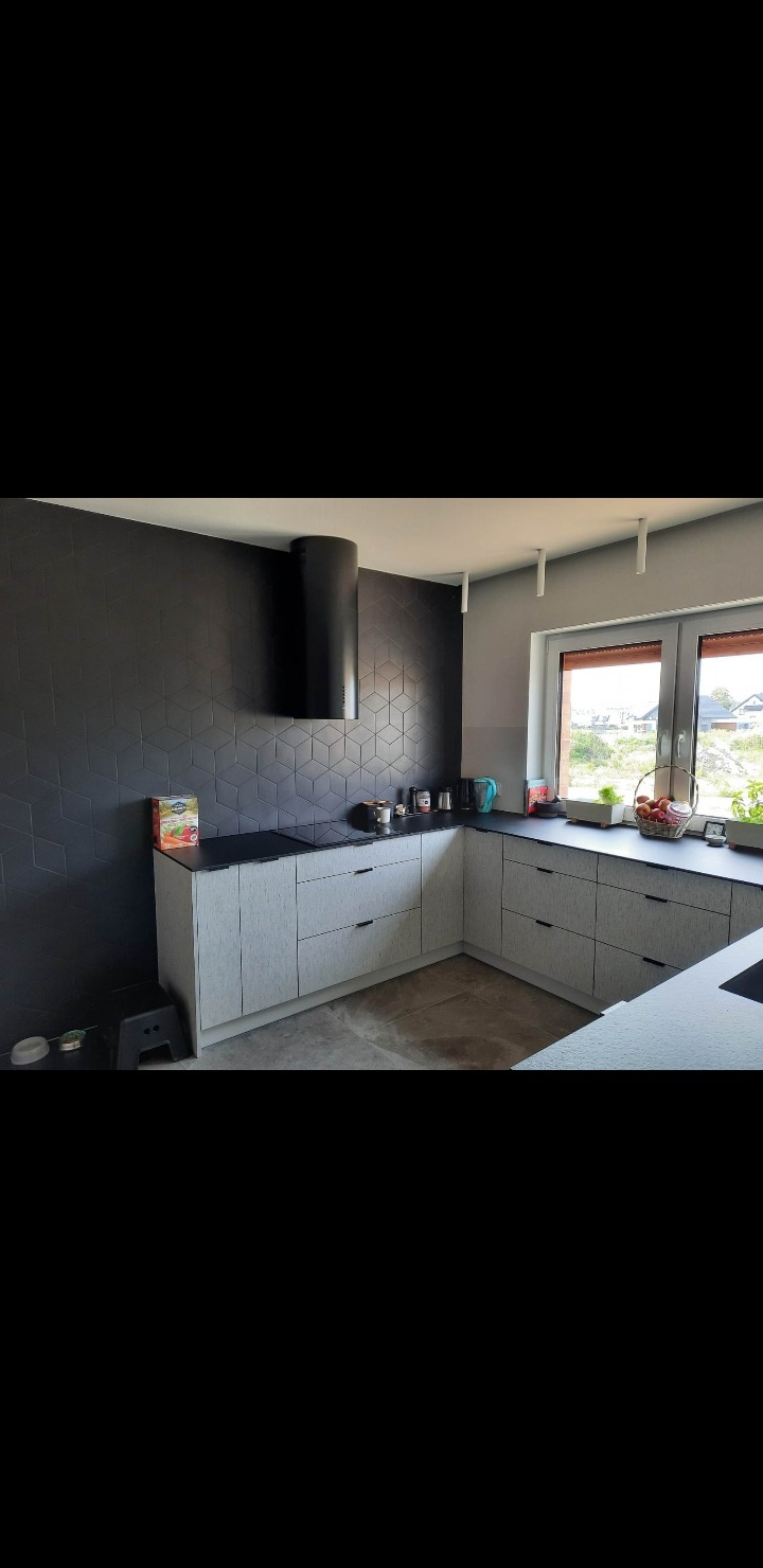 Nowoczesna kuchnia z szarymi szafkami i czarną ścianą z geometrycznym wzorem. Widoczny okap cylindryczny i okno z widokiem na zieleń.