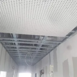 Montaż sufitu podwieszanego z perforowanej płyty w korytarzu, widoczne metalowe profile i instalacje nad sufitem, ściany w trakcie szpachlowania.