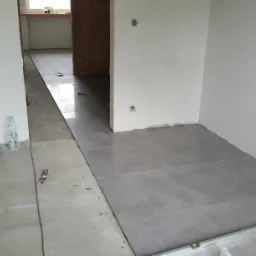 W trakcie układania szarych płytek podłogowych w pomieszczeniu, widoczne narzędzia glazurnicze, paczka płytek, poziomica, szczotka i butelka wody.