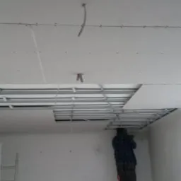 Montaż sufitu podwieszanego: pracownicy na drabinach instalują konstrukcję metalową i płyty gipsowo-kartonowe, widoczne przewody elektryczne i oświetlenie punktowe w trakcie instalacji.