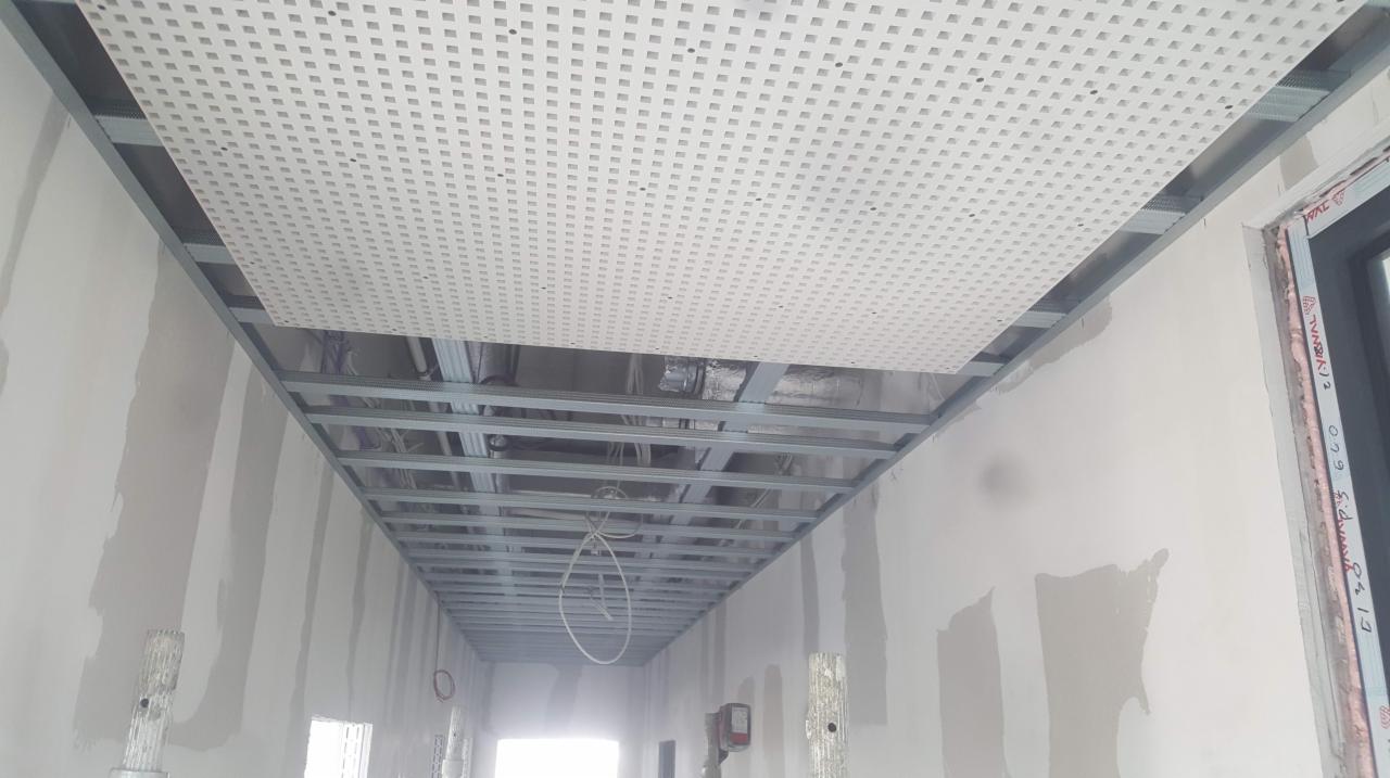 Montaż sufitu podwieszanego z perforowanej płyty w korytarzu, widoczne metalowe profile i instalacje nad sufitem, ściany w trakcie szpachlowania.