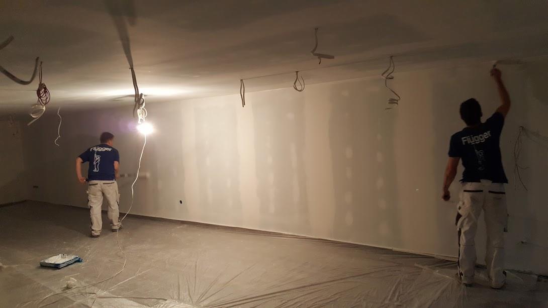 Dwóch malarzy w koszulkach Flűgger szpachluje ściany w dużym pokoju z odsłoniętymi przewodami, podłoga zabezpieczona folią.