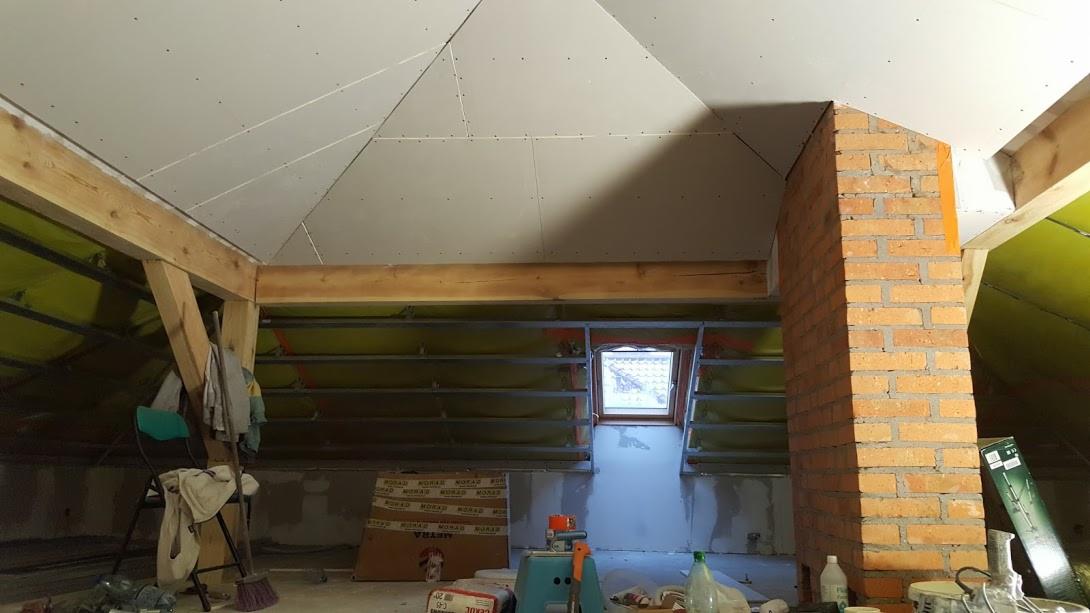 Poddasze w trakcie wykańczania: widoczne ocieplenie, konstrukcja drewniana, komin z cegły oraz płyty kartonowo-gipsowe na skośnym suficie, przygotowane do szpachlowania.