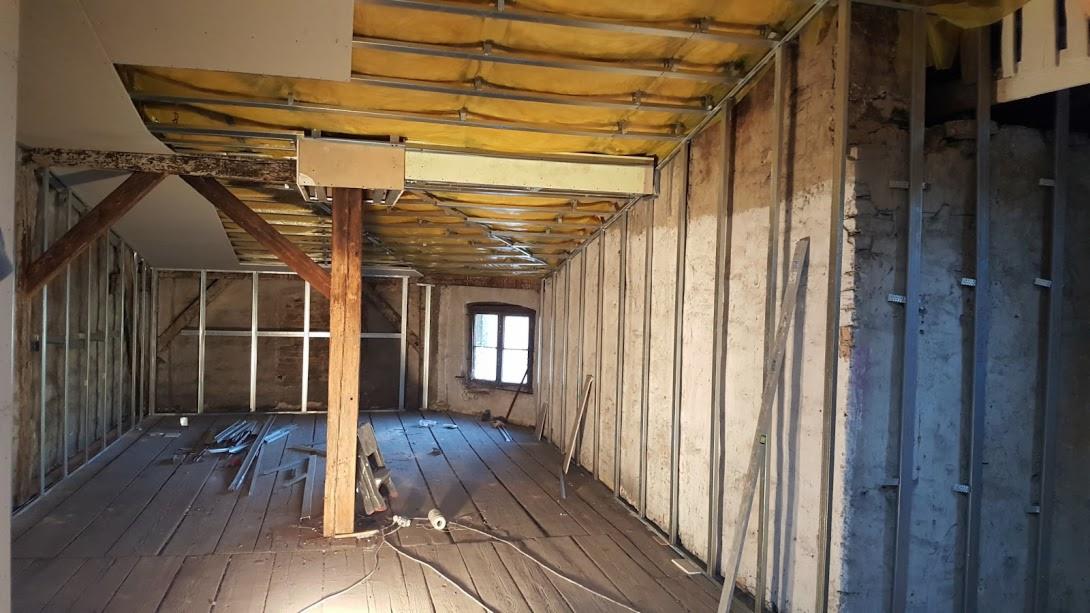 Wnętrze w trakcie remontu: surowe drewniane podłogi, metalowa konstrukcja ścian z widoczną starą cegłą, izolacja stropu z żółtą wełną mineralną, drewniane belki stropowe i okno w tle.
