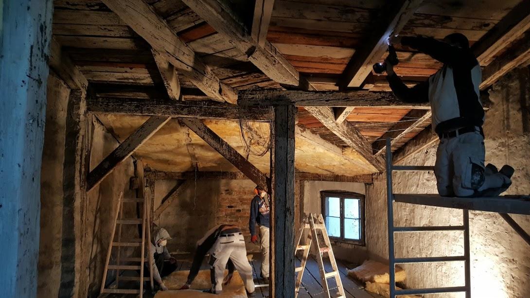 Renowacja poddasza w starym domu: pracownicy na rusztowaniach i drabinach wykonują prace wykończeniowe przy drewnianej konstrukcji dachu, widoczne ocieplenie i fragment okna.