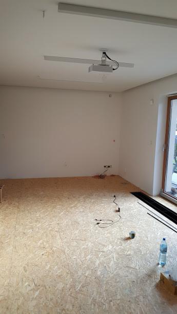 Surowa płyta OSB na podłodze w trakcie remontu, widoczne kable elektryczne, butelka wody i listwy przypodłogowe przy oknie.