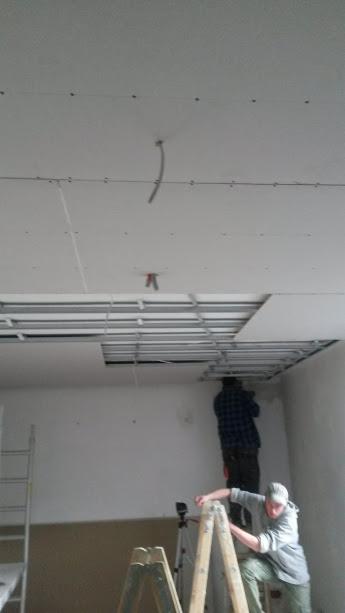 Montaż sufitu podwieszanego: pracownicy na drabinach instalują konstrukcję metalową i płyty gipsowo-kartonowe, widoczne przewody elektryczne i oświetlenie punktowe w trakcie instalacji.