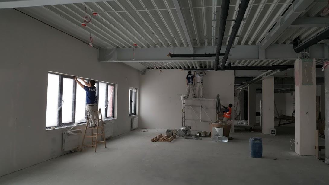 Wnętrze w trakcie remontu: pracownicy na rusztowaniu i drabinie przygotowują ściany do malowania, okna zabezpieczone folią, widoczne puszki z farbą i narzędzia.