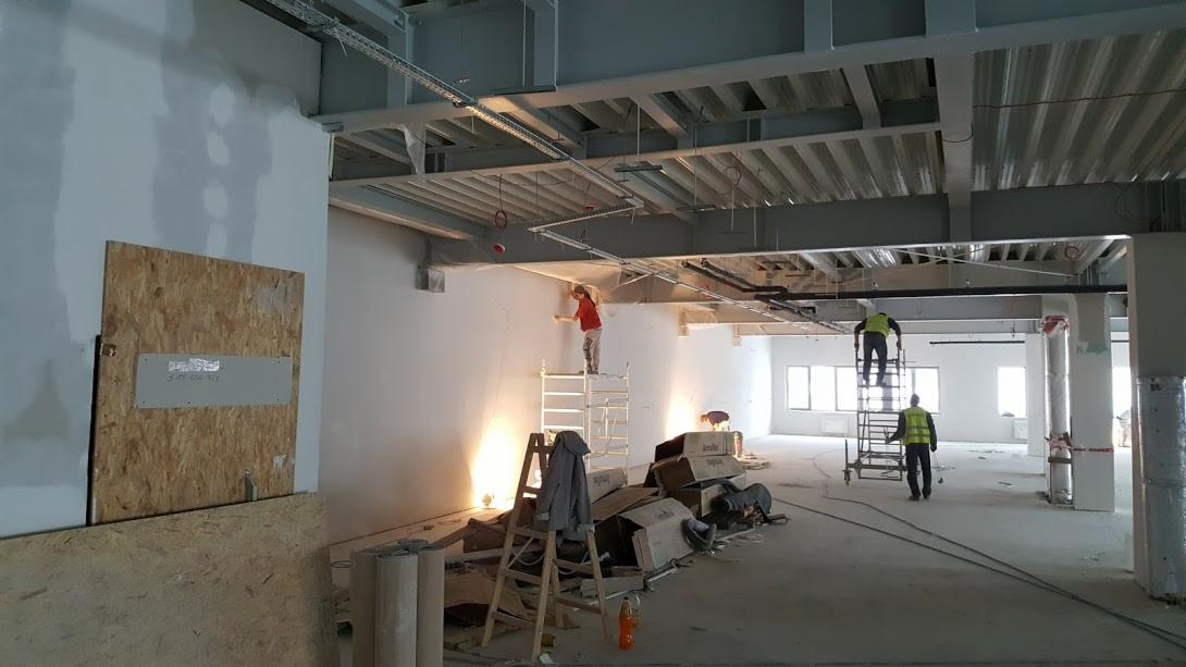 Wnętrze w trakcie prac wykończeniowych: pracownicy na rusztowaniach, szpachlowanie ścian w dużym pomieszczeniu z surowym stropem i instalacjami pod sufitem, widoczne materiały budowlane i narzędzia...