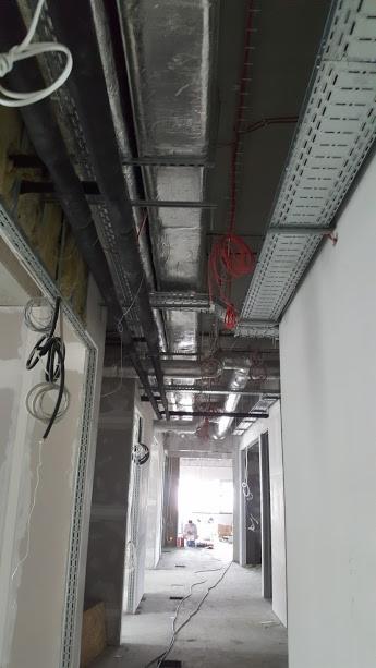 Korytarz w stanie surowym z widocznymi instalacjami pod sufitem: rury wentylacyjne, kable elektryczne w korytkach, przewody, ściany przygotowane do szpachlowania.