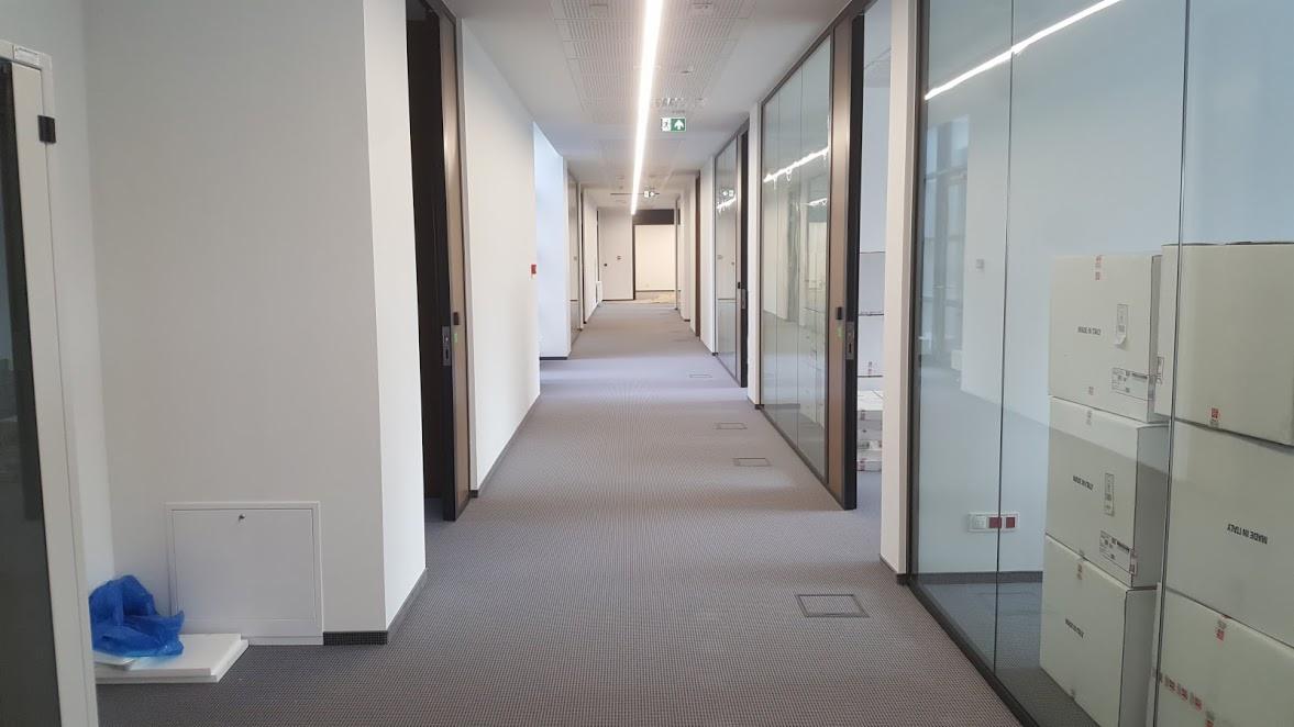 Długi korytarz biurowy z szarą wykładziną, białymi ścianami, rzędem drzwi i szklanych ścianek działowych, przy których stoją kartony. Widoczne oświetlenie liniowe LED i niebieska folia na podłodze...