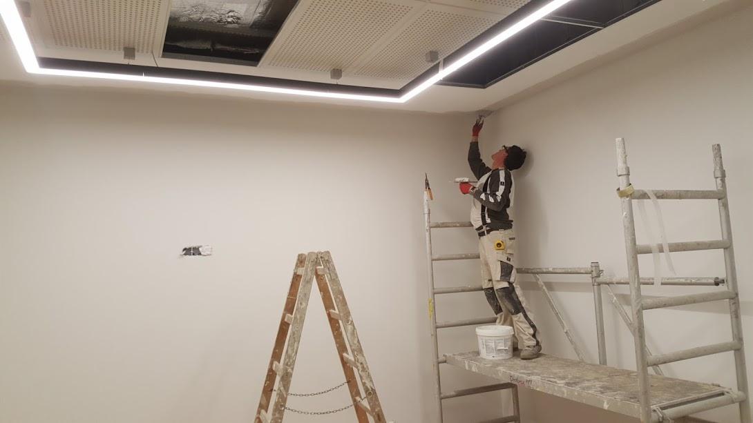 Pracownik na rusztowaniu maluje ścianę w pomieszczeniu z podwieszanym sufitem i oświetleniem liniowym LED, widoczne dwie drabiny i wiadro z farbą.